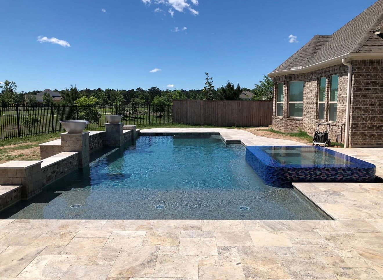 Modern rectangular pool with blue glass mosaic spa, fire bowls on stone pillars, and travertine deck — Houston, TX