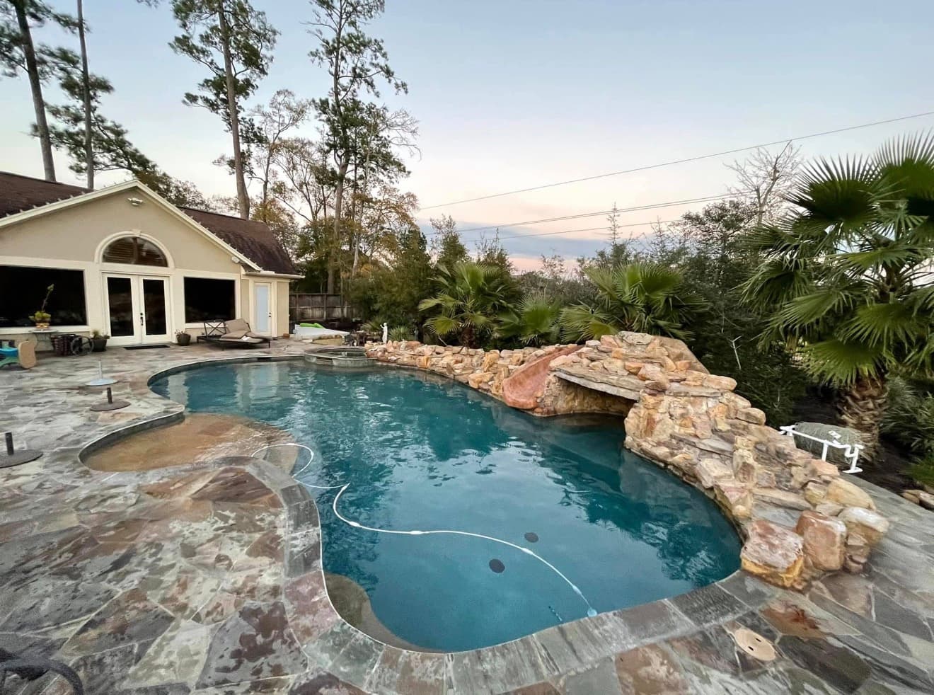 Resort-style freeform pool at dusk with rock waterfall slide, raised spa, and palm tree landscaping — Houston, TX