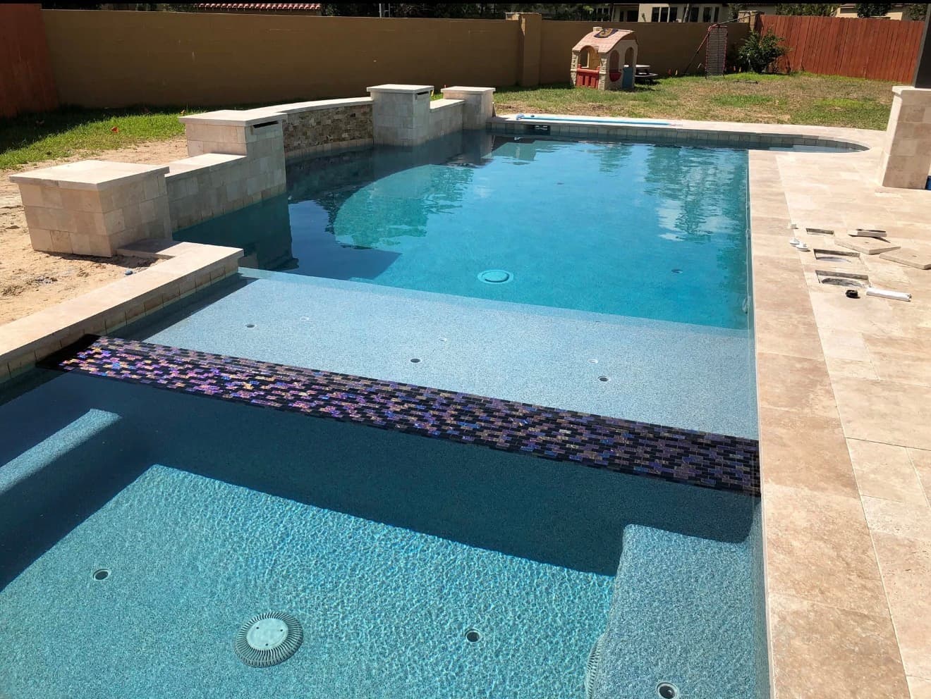 Freeform pool under construction with curving travertine paver deck and raised stone scupper wall feeding twin spouts, covered patio and outdoor kitchen behind — Houston, TX